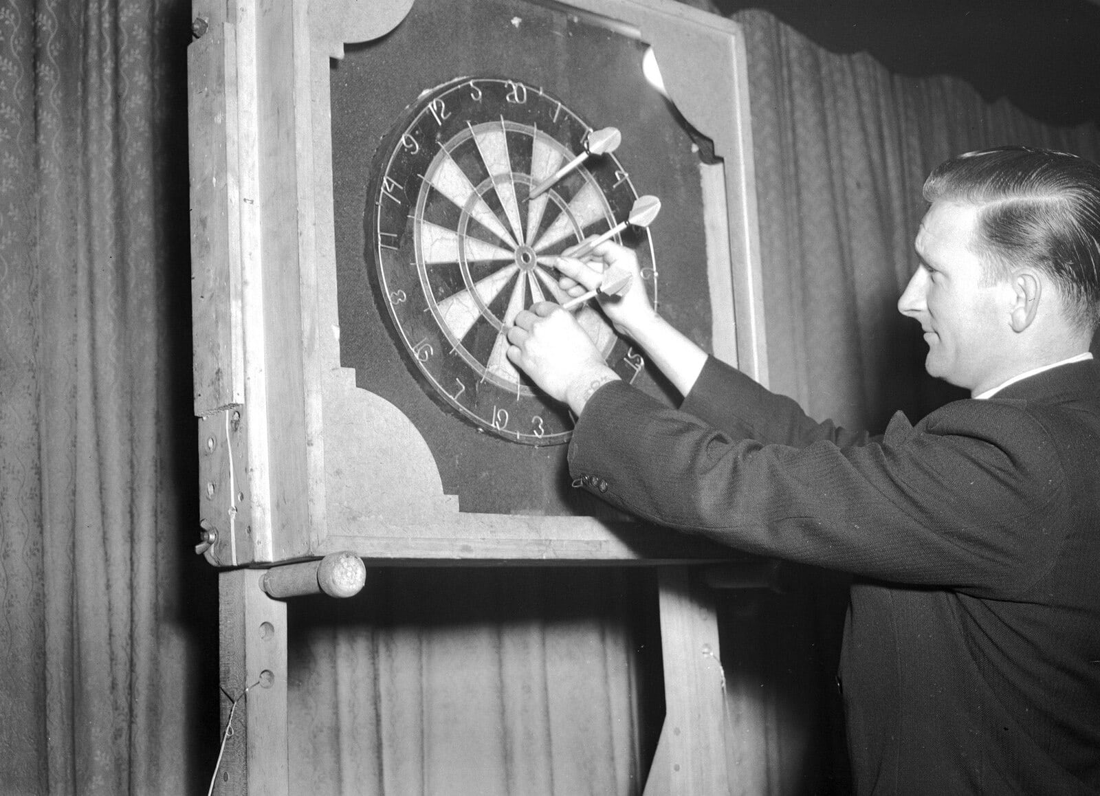 Black and white photo of a man in a suit retrieving darts from a vintage dartboard in a wooden frame.