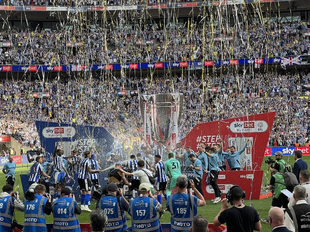 Sheffield Wednesday players celebrate promotion at Wembley Stadium with confetti and the Sky Bet League One playoff trophy, surrounded by cheering fans and photographers.