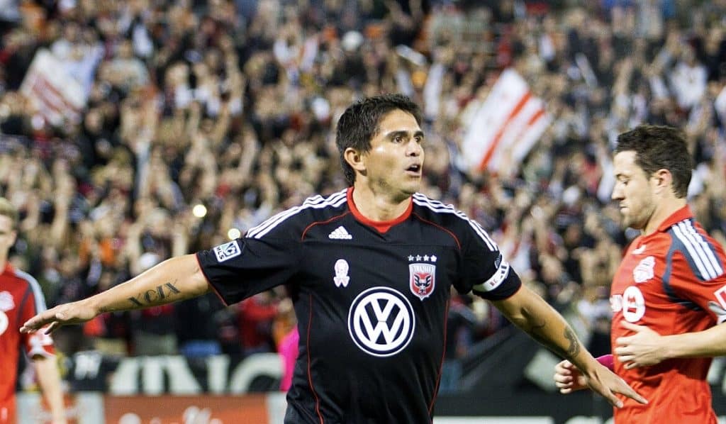 A legendary MLS player from D.C. United celebrates after scoring a goal during a match, with Toronto FC players and a cheering crowd in the background.